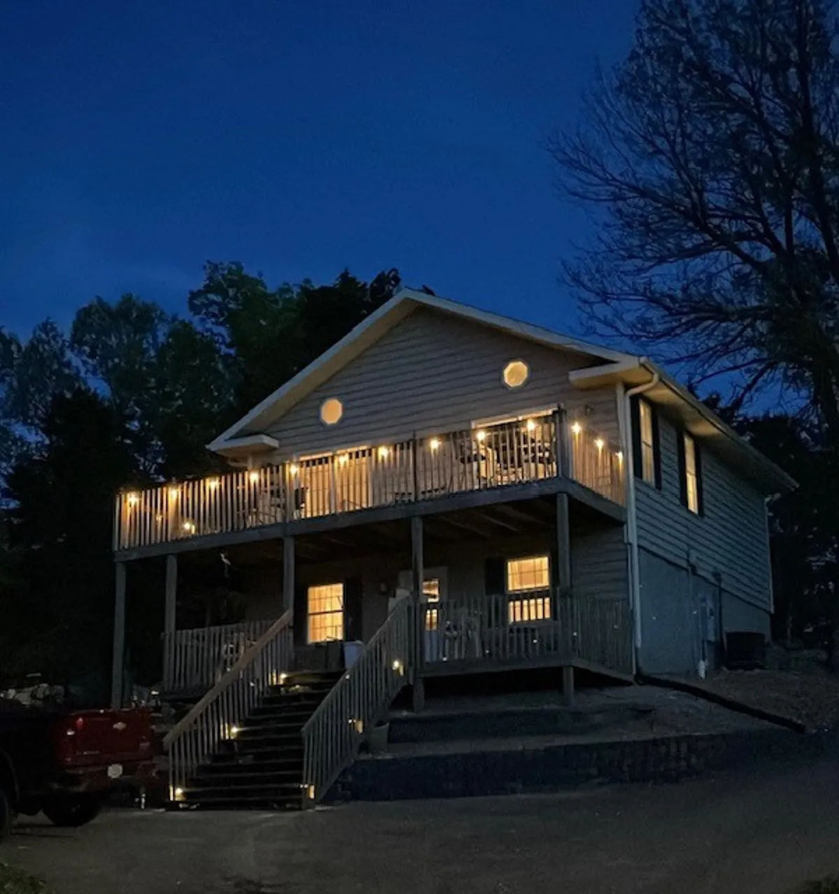 An inviting exterior shot featuring a warmly illuminated porch, decorative lights along the railing, and a welcoming glow emanating from the home's windows. The scene suggests a lively and comfortable space for evening gatherings.