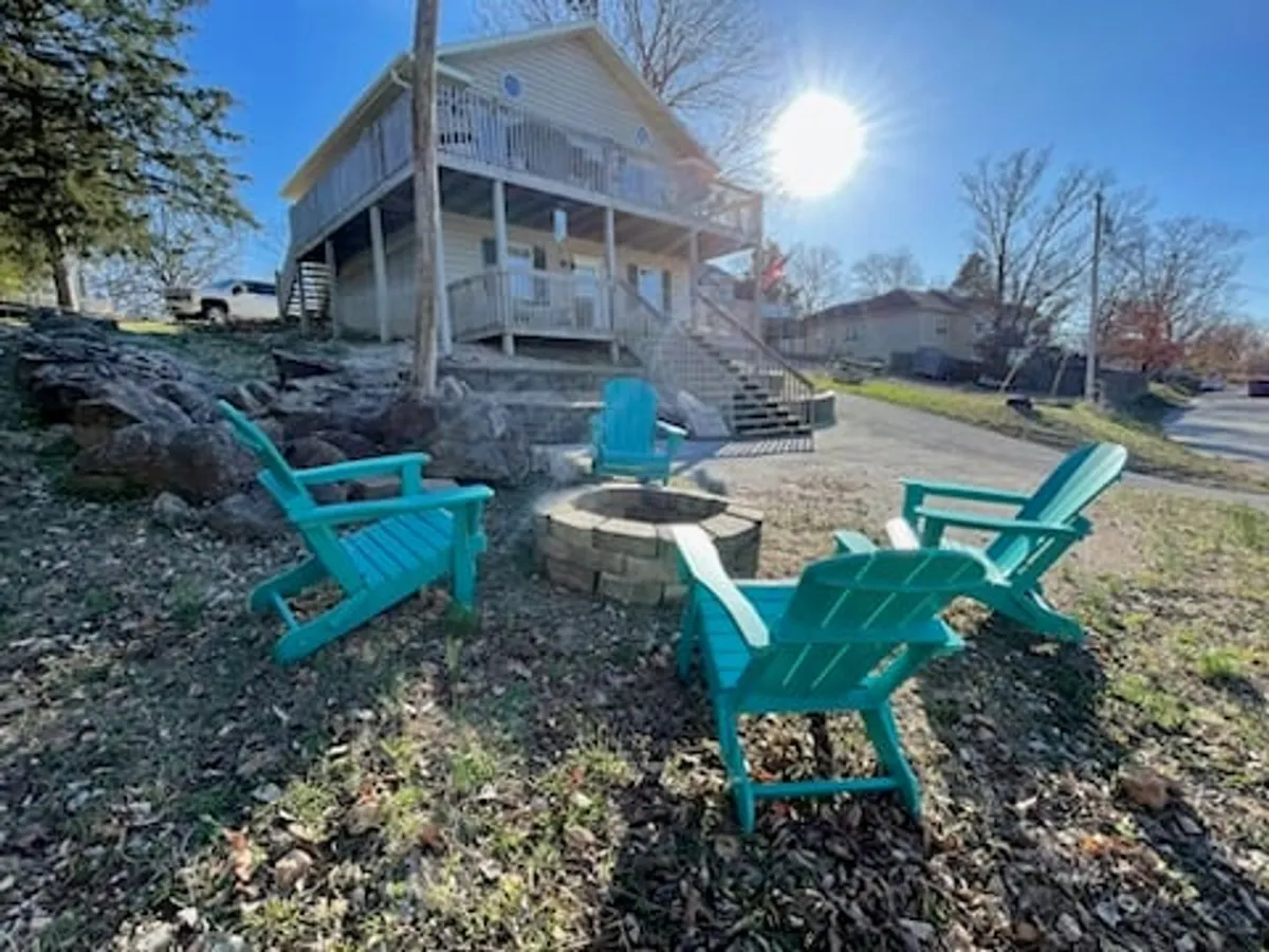 A relaxed outdoor setup with turquoise Adirondack chairs encircling a fire pit on a deck, accompanied by the house and a backdrop of natural rock formations. The scene exudes sociable energy and a casual, festive mood.