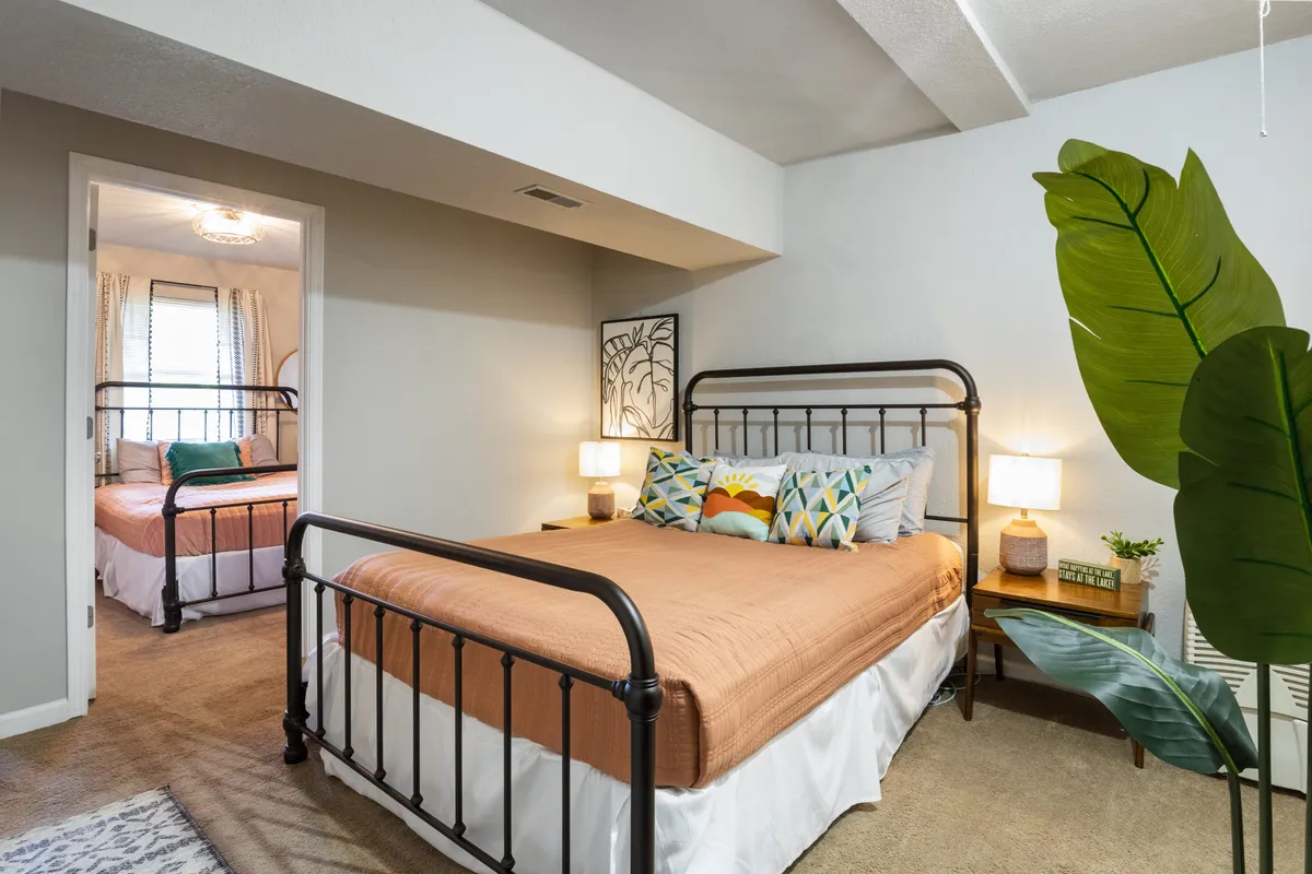 A welcoming lower level sleeping area featuring two metal-framed beds, decorative pillows, green plant accents, artwork on the wall, and nightstands with lamps. The setup feels cozy and flexible, ideal for accommodating groups or families.