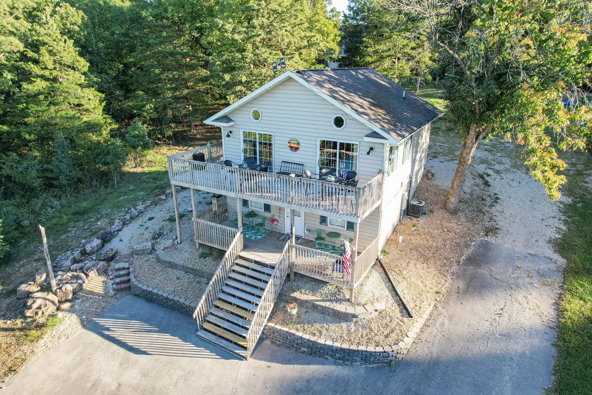 A spacious wooden deck stretches out from the home, surrounded by tall trees and featuring large front steps and an American flag. The area is set up for gatherings, with plenty of room for outdoor seating and activities. The greenery and natural setting create a relaxing, inviting atmosphere.