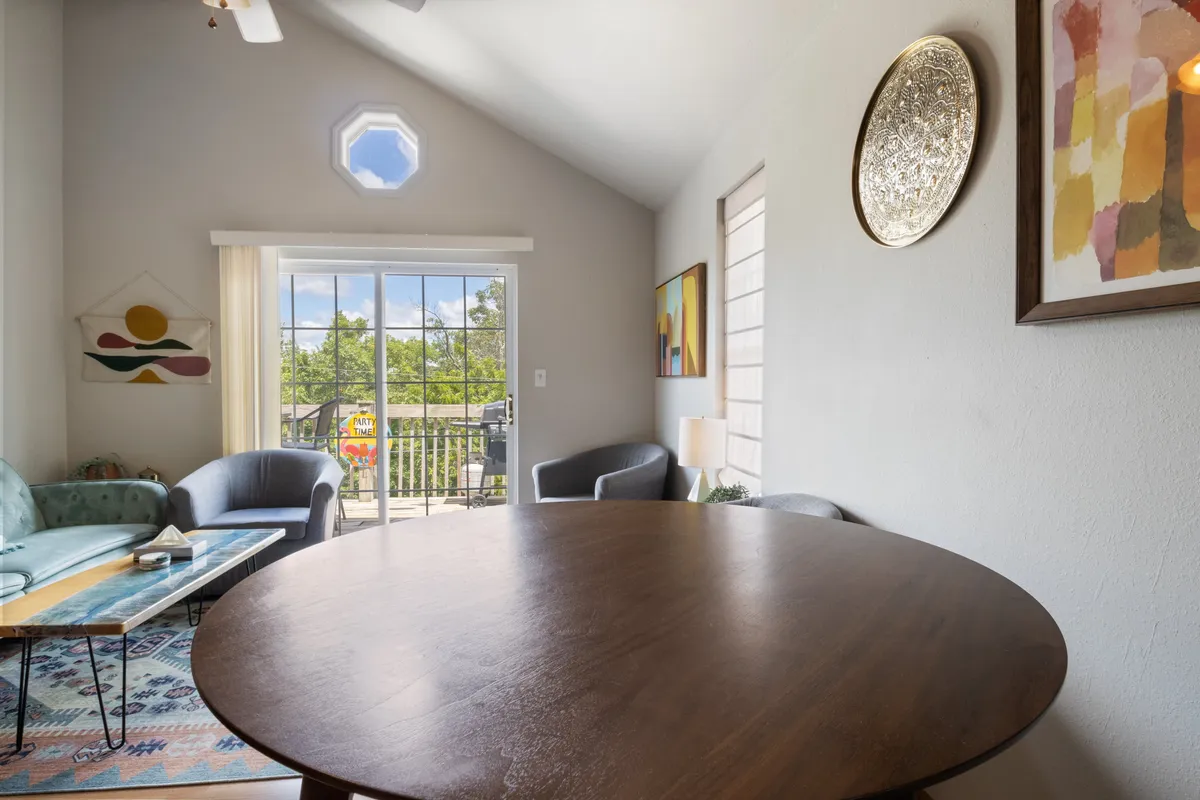 A stylish, open-plan living and dining area with a round wooden table, blue sofa, and expansive glass doors leading to a balcony. Colorful artwork adorns the walls, while sunlight streams in, creating a welcoming, creative atmosphere.