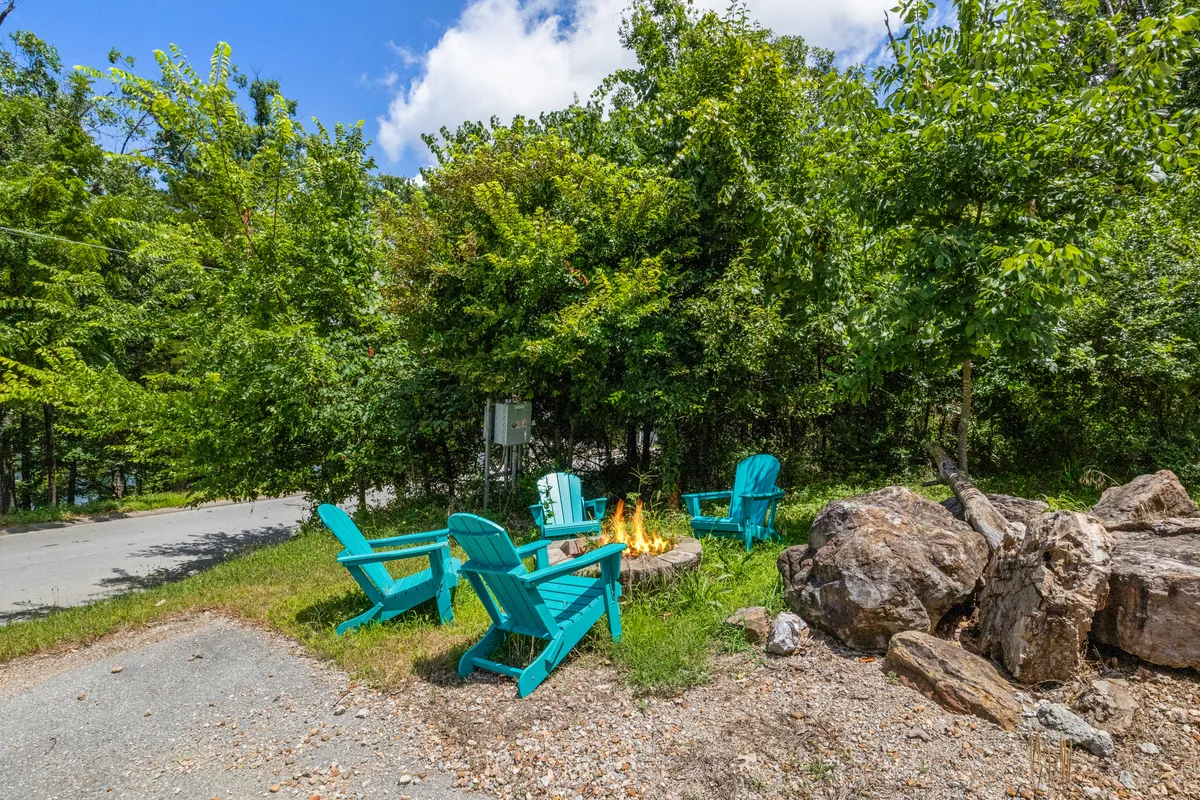 A cozy fire pit area encircled by bright blue Adirondack chairs, set amidst large rocks and rich greenery. The inviting setting suggests warm evenings, storytelling, and shared moments under the stars.