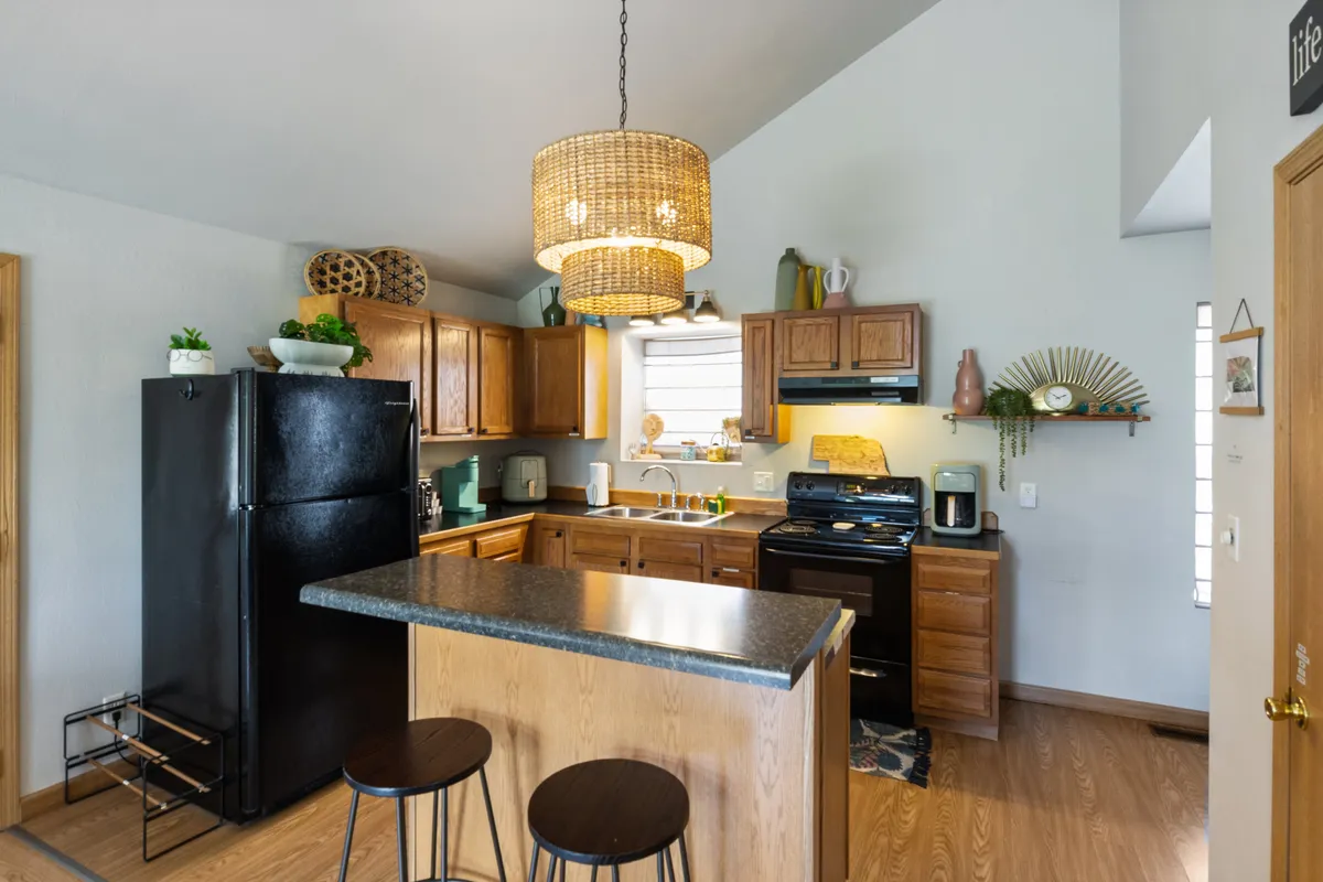 A kitchen scene anchored by an island counter with stools, a modern hanging light fixture, and wooden cabinetry. The space is styled for both meal prep and casual conversation, with enough room for multiple people to gather.