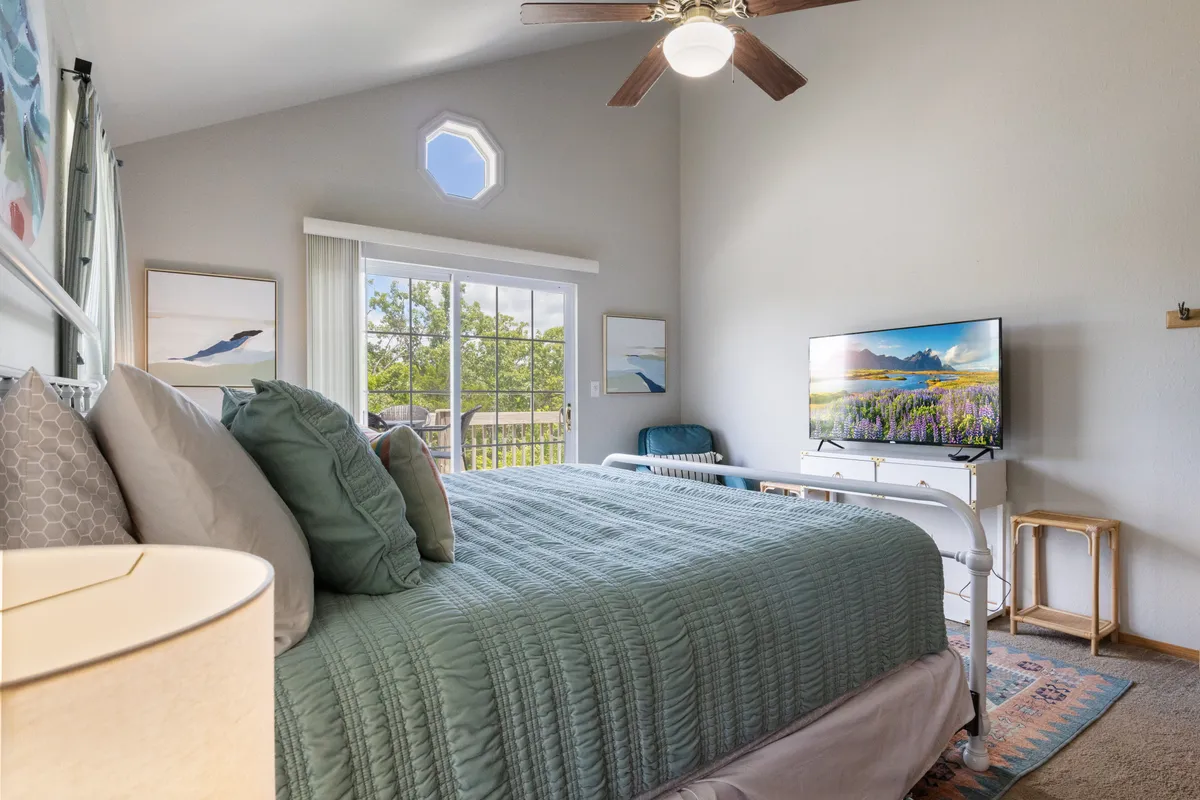 A tranquil master bedroom featuring a bed with teal bedding, a large window that allows in natural light, a TV displaying a serene landscape, and tasteful artwork on the walls. The room has a peaceful, inviting ambiance, perfect for restful nights.