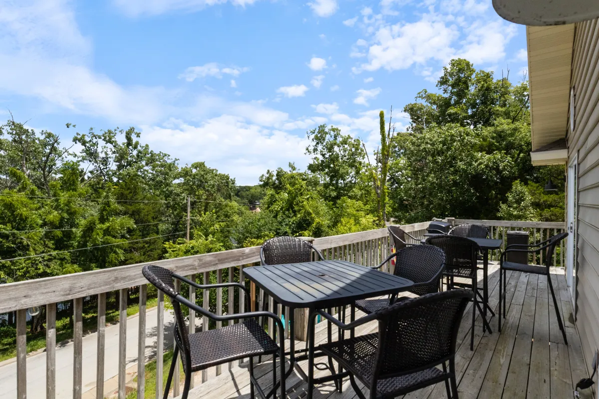 A tranquil wooden deck with black patio furniture overlooks lush greenery and a clear sky. The setup is ideal for outdoor dining or simply relaxing with friends and family. The inviting space captures the transition between indoor and outdoor living.
