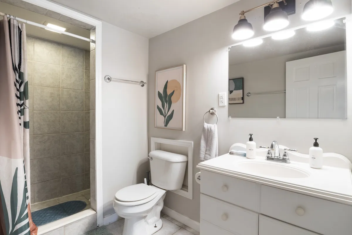 A modern bathroom with a vanity and sink, a spacious shower area with a glass door, and tasteful artwork on the wall. The space is clean, calm, and well-lit, suggesting comfort and convenience.
